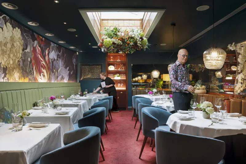 Elegant Pied à Terre dining room with floral wall art, skylight, hanging plants, set tables, and staff in service.