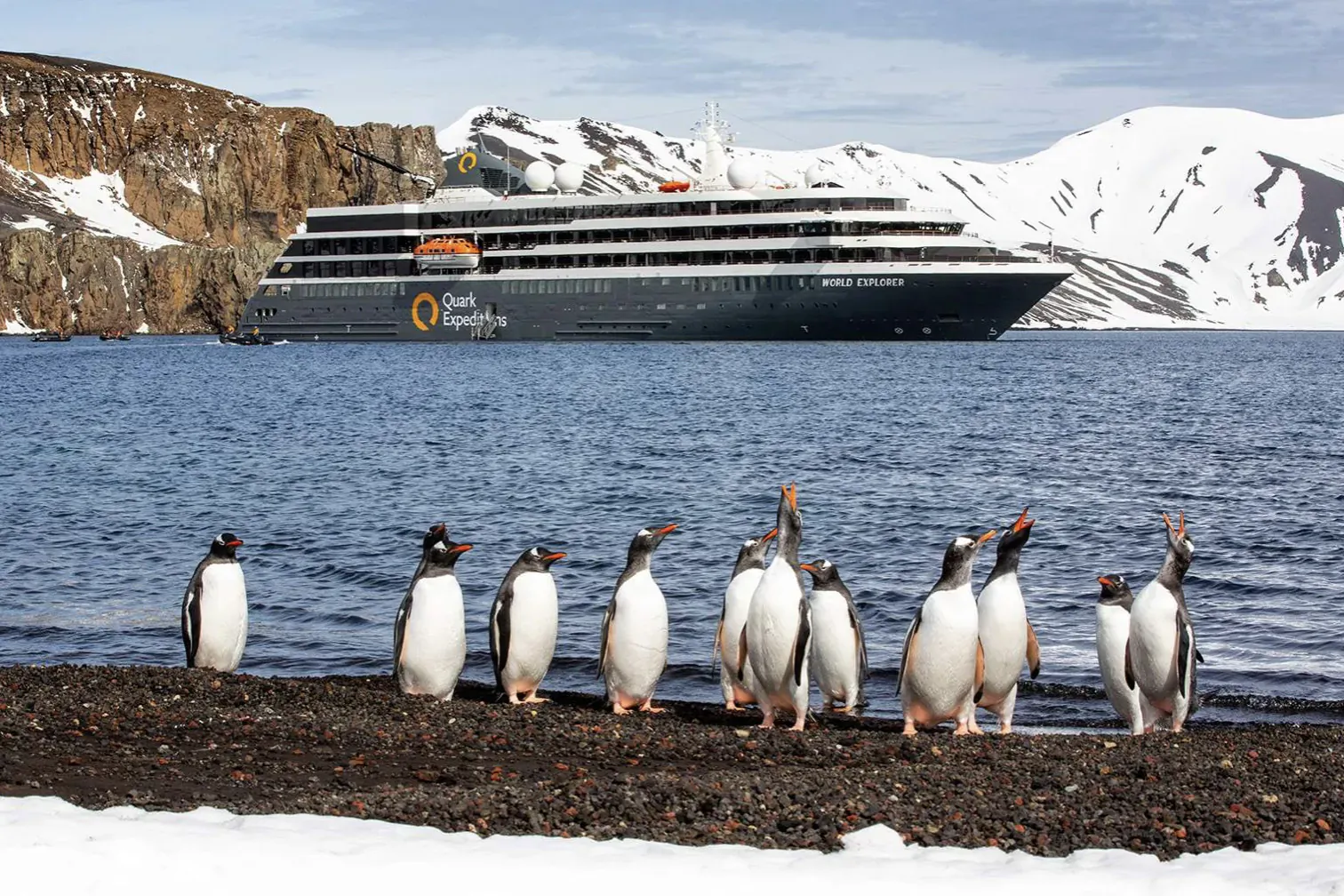 Group of gentoo penguins on snowy shore with luxury cruise ship in Antarctic bay, snow-capped mountains behind.