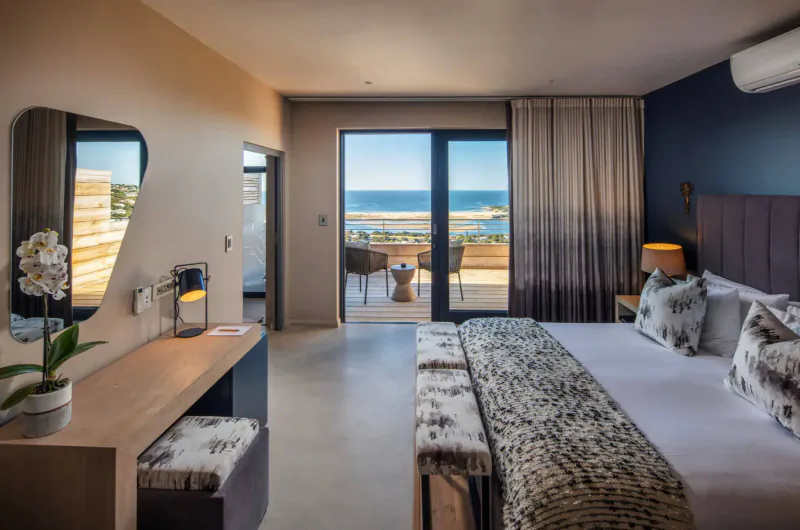Luxurious hotel bedroom with king bed, vanity, orchids, and balcony overlooking ocean.