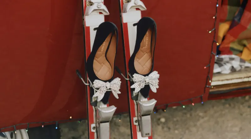 Black Kurt Geiger high-heeled shoes with white bows on white skis beside red holiday-decorated van