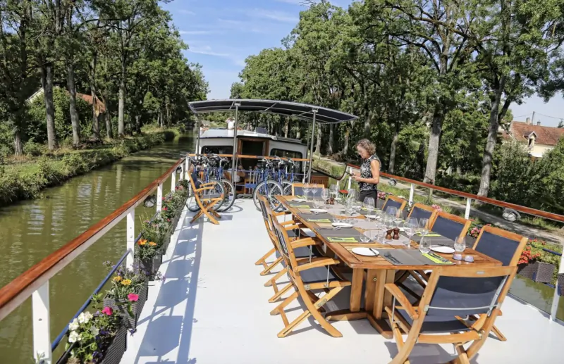 Woman standing by set dining table with wine glasses on a houseboat deck along tree-lined Burgundy canal