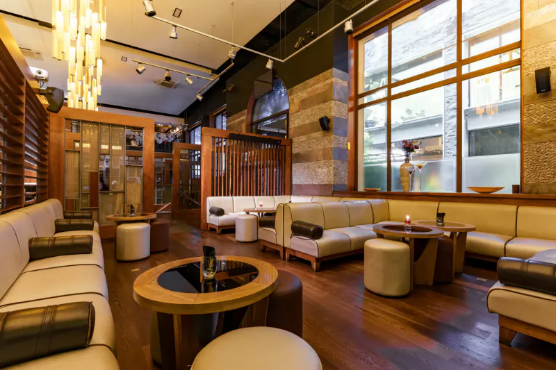 Elegant Mint Leaf London gallery lounge with wooden decor, cream sofas, poufs, round tables, and pendant lights.