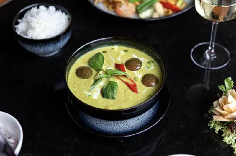 Bowl of green Thai curry with basil, peppers, and meatballs, served with rice, garnished with edible flowers and wine glass.