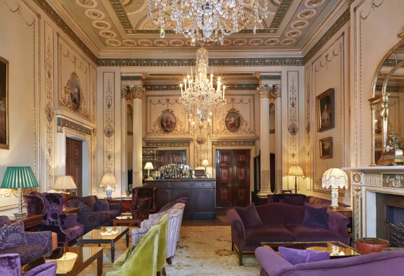 Opulent Home House lounge with grand chandelier, ornate ceilings, purple velvet sofas, bar, lamps, and fireplace.