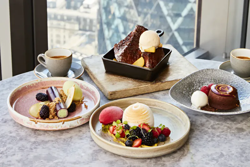 Assortment of desserts on plates at Duck and Waffle: chocolate cake with ice cream, crepe cake, pavlova with berries, creme brulee, city skyline view.