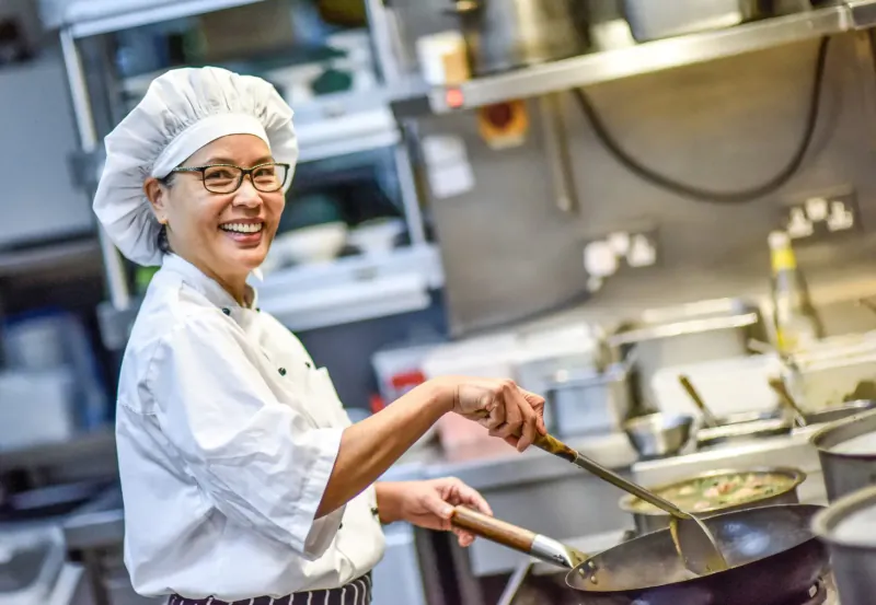 Smiling Asian female chef in white uniform and hat stir-frying vegetables in a wok in professional kitchen