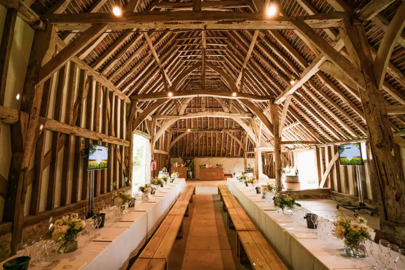 Spacious rustic barn interior with long wooden tables set for dining, flowers, and screens, warm lighting from beamed ceiling.