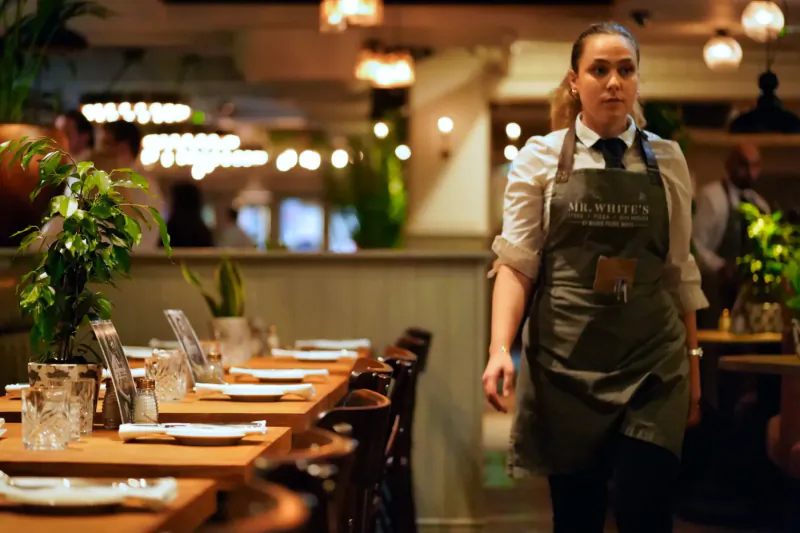 Server in Marco Pierre White apron walks past wooden dining tables with plants in warm restaurant lighting