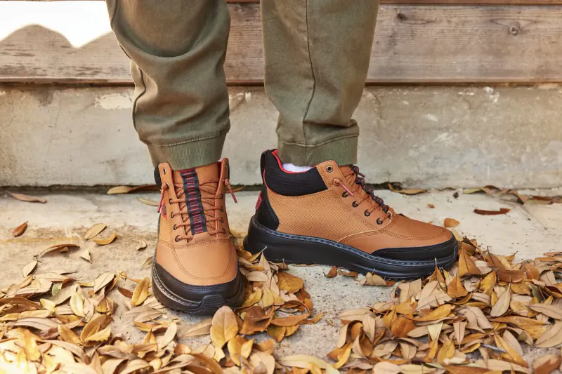 Close-up of brown and black laced boots on khaki pants, standing on autumn leaves against wooden backdrop.