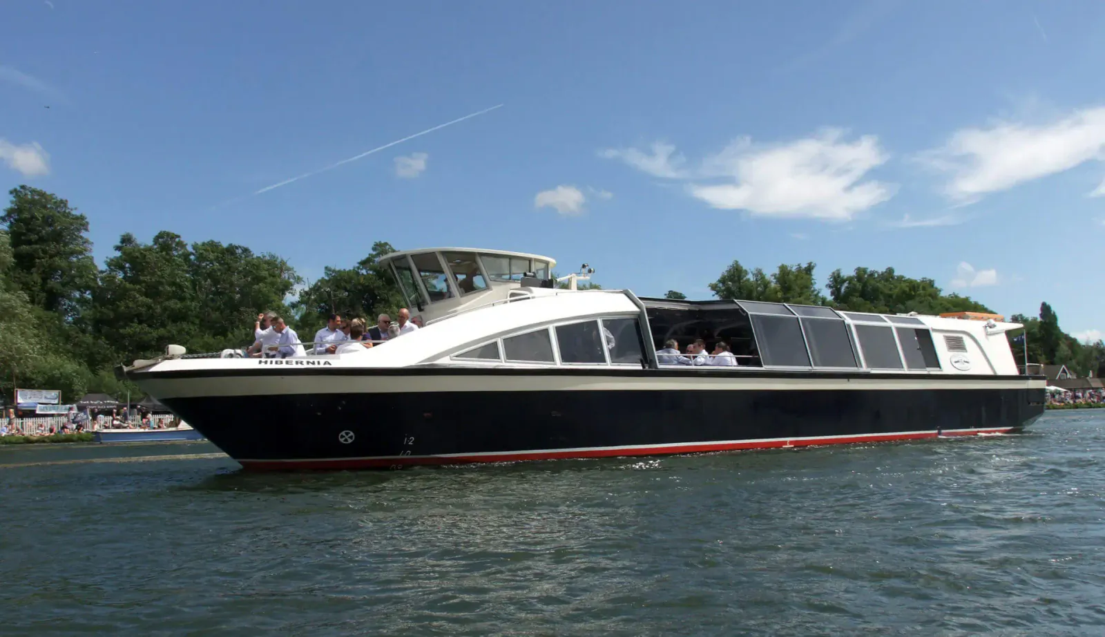 White and black Hobbs of Henley tour boat with passengers cruising on river, lined by trees under blue sky.