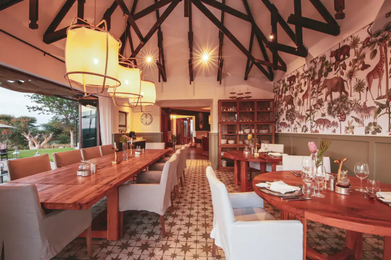 Elegant Namibian dining room with wooden beams, pendant lamps, animal print walls, long wooden table, and garden view.