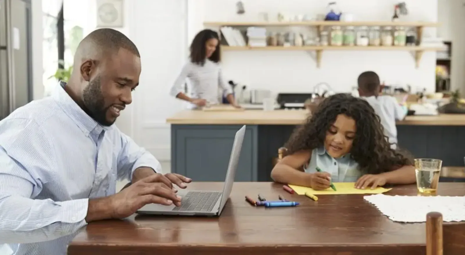Black father working on laptop at kitchen table while daughter draws nearby, mother cooks in modern kitchen.