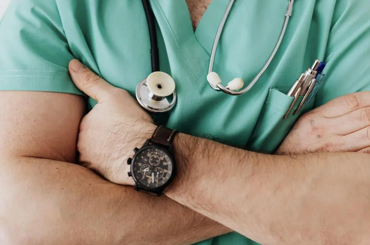 Male doctor in green scrubs and stethoscope, arms crossed, wearing watch and pen in pocket
