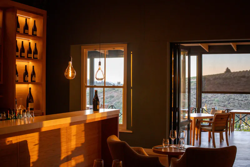 Cozy bar at Brookdale Wine Estate with wooden shelves of wine bottles, pendant light, sunset view over hills and patio seating.