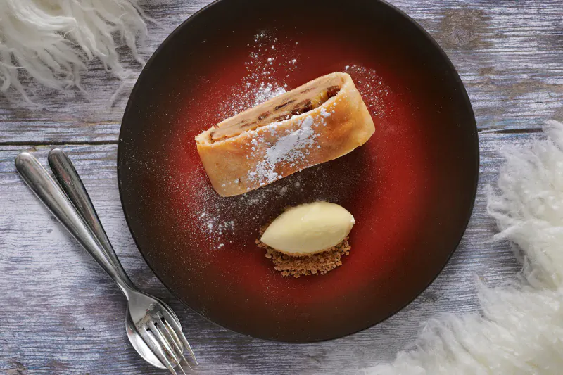 Apple strudel with powdered sugar, crème fraîche quenelle, and crumbs on red plate, fork and knife, snowy fur backdrop.