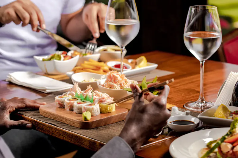 Close-up of diners' hands using utensils to eat sushi rolls, shrimp, fries, and salads at Polo Club table with wine glasses.