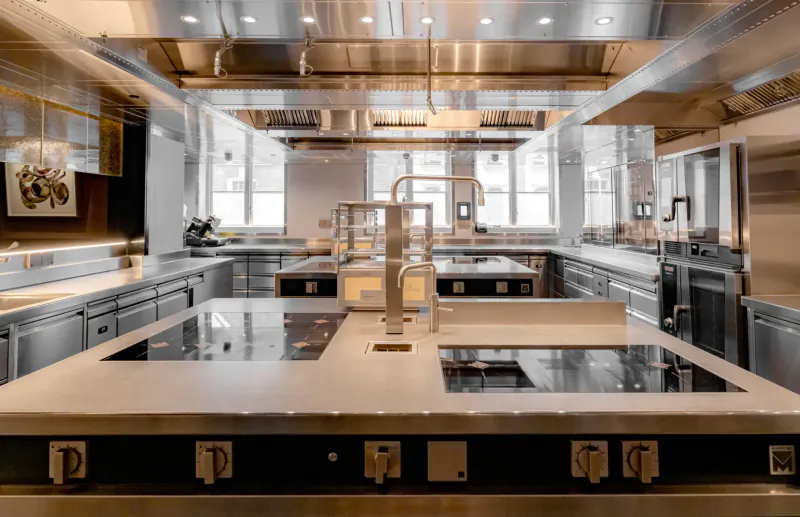 Modern professional kitchen with stainless steel counters, induction cooktops, central faucet, and overhead ventilation.