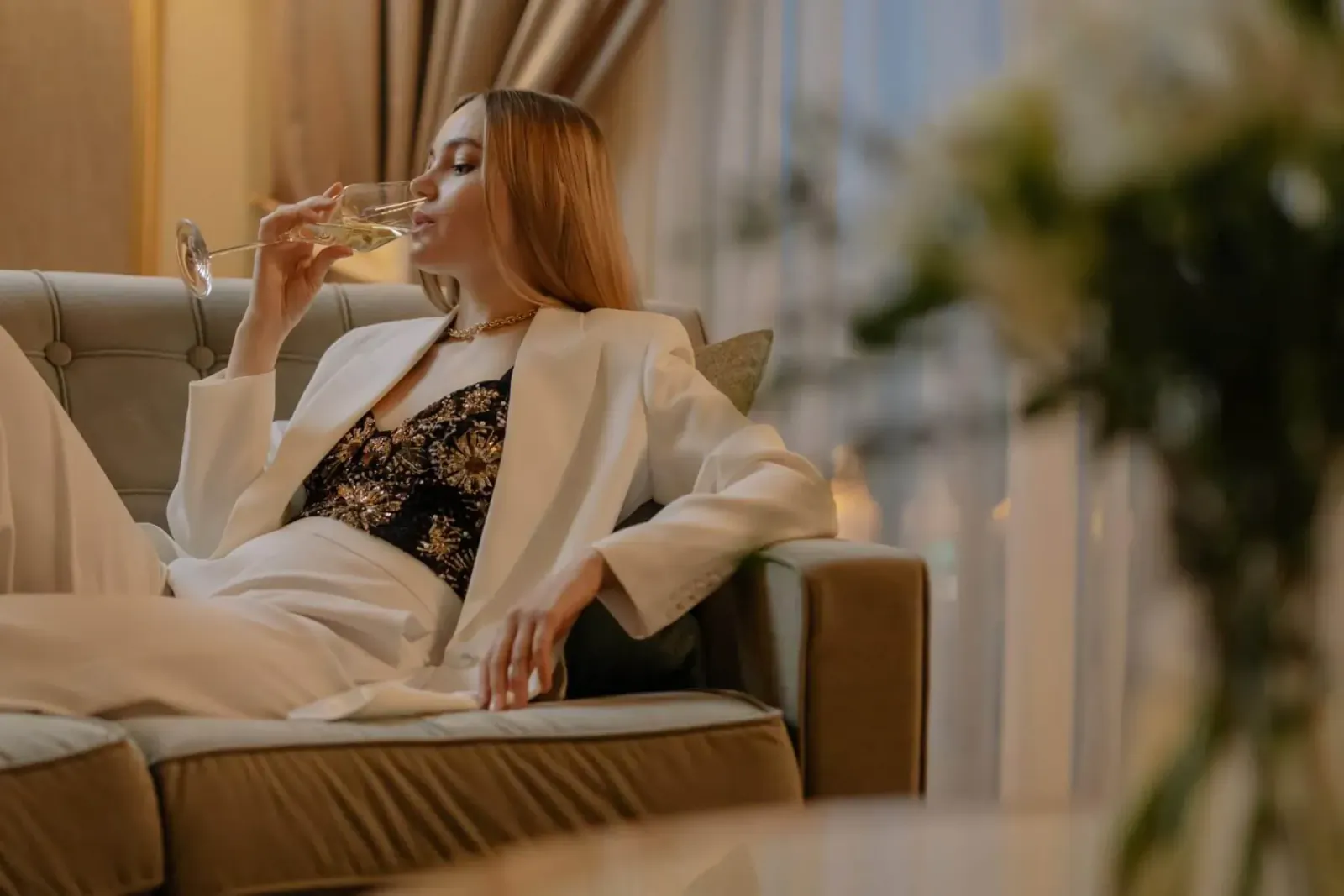 Blonde woman in white blazer and pants sips champagne on luxury couch amid flowers and candles