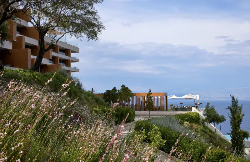 Angsana Corfu Resort & Spa on a lush hillside overlooking the sea, with orange buildings, pool, and umbrella.