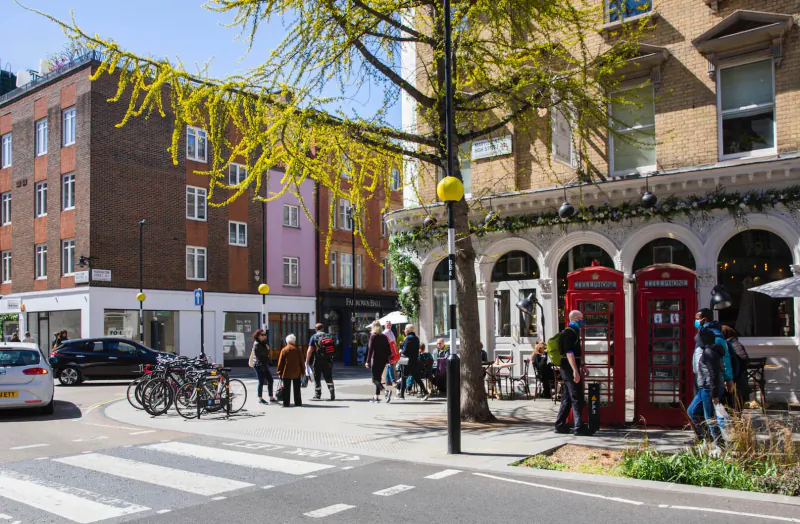 Marylebone Village street scene: red phone booths, yellow lamp post, pedestrians, bikes, cars at zebra crossing under golden tree.