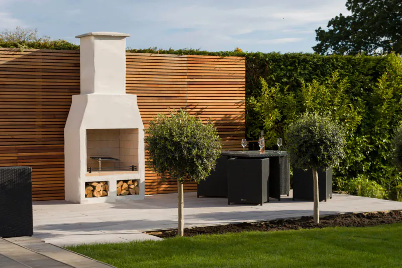 Schiedel Isokern Volcanic white garden fireplace with firewood, flanked by wooden slat fence, hedges, olive trees, and black outdoor table with wine glasses on patio