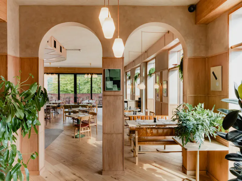 Sophisticated dining area at The Broadcaster, London, with wooden tables, chairs, potted plants, arches, and panoramic garden views.