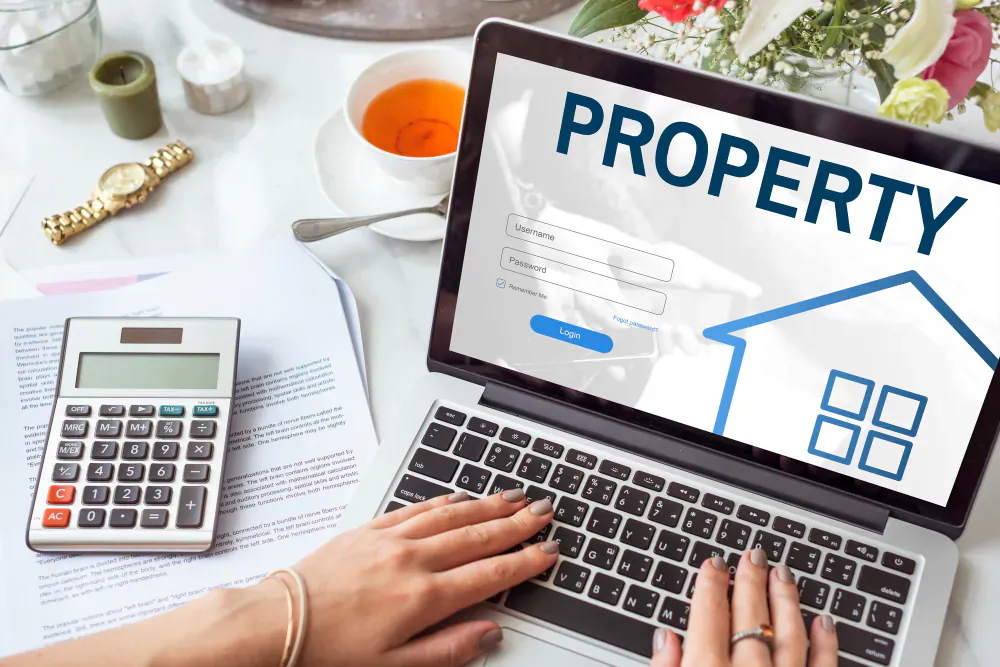 Woman's hands typing on laptop showing 'PROPERTY' login with house icon, calculator, papers, tea on desk.