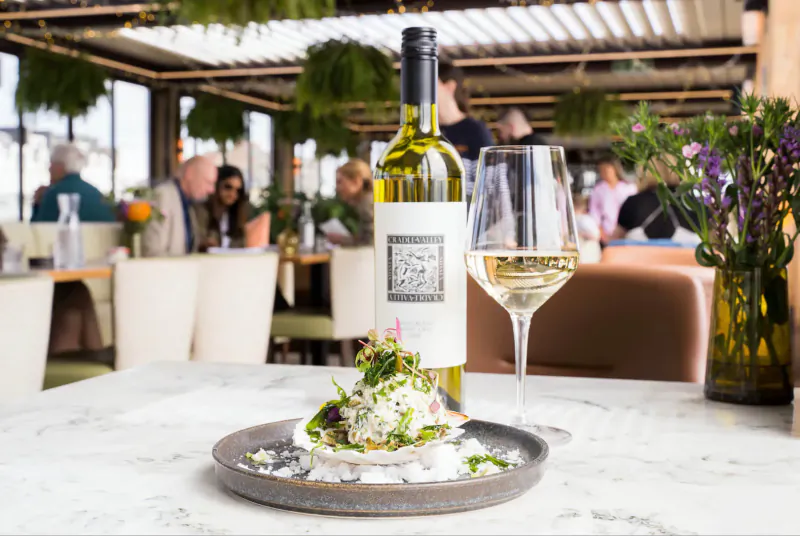 Close-up of white wine bottle and glass beside seafood dish on table in sunny outdoor restaurant with blurred patrons