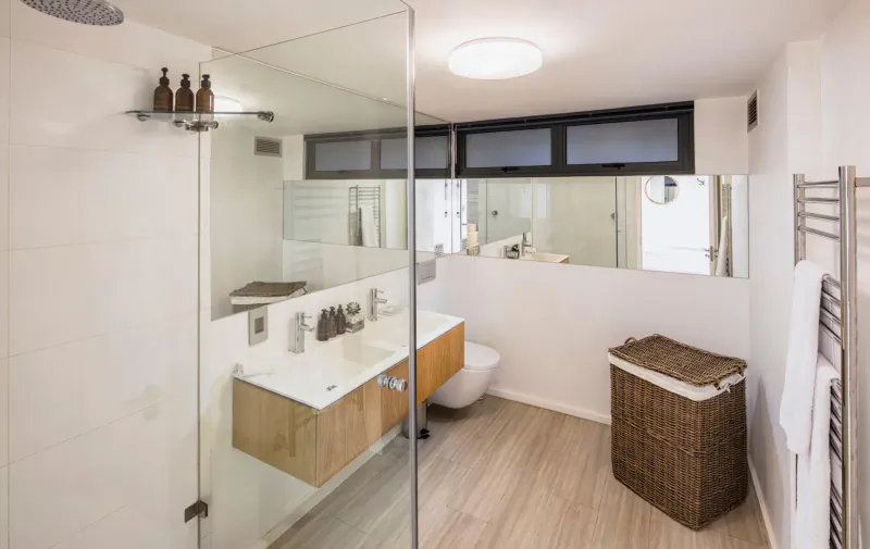 Modern white bathroom with glass shower, wall-mounted sink, toilet, towel rack, and wicker laundry basket at Spanish Farm Guest Lodge.