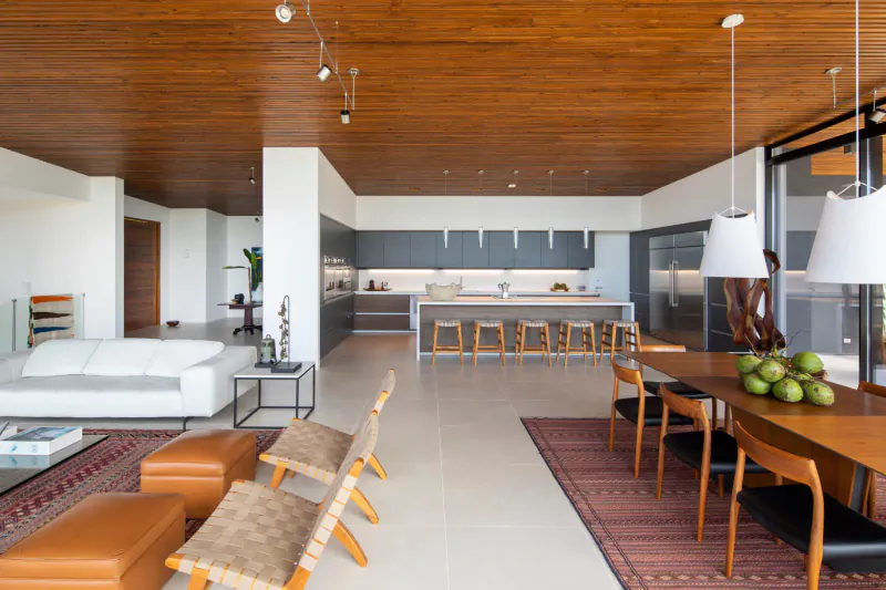 Spacious modern luxury living room with wooden ceiling, white kitchen, dining table with green apples, and rattan chairs.