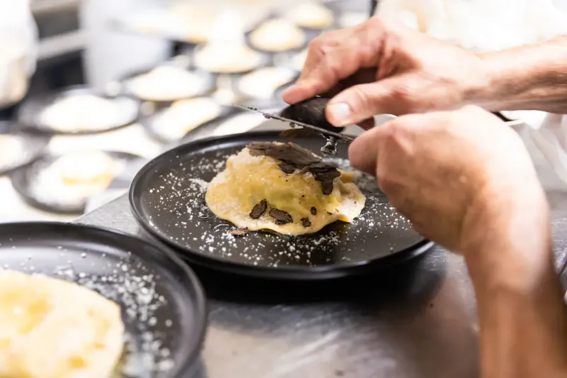 Chef's hands spreading chocolate onto blini topped with caviar on black plate in kitchen, surrounded by more blinis.