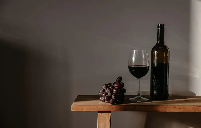 Glass of red wine, bottle, and cluster of grapes on wooden table in soft light