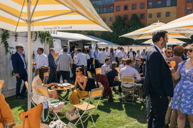 Elegant crowd in summer dresses and suits mingling at Veuve Clicquot tents on green lawn at concours d'elegance event