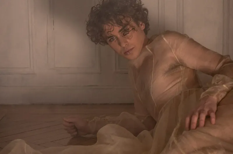 Javi Costa Polo with curly hair reclines on wooden floor in sheer beige gown, soft lit room with paneled walls.