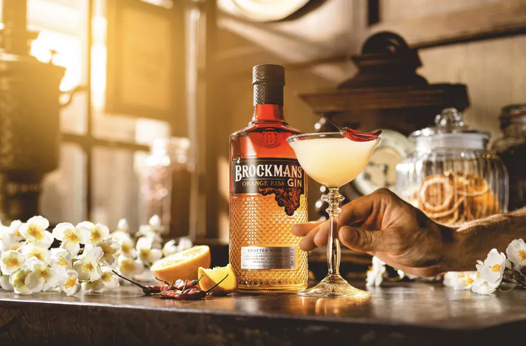 Brockmans Gin bottle and Orange Kiss cocktail in martini glass held by hand, with orange slices, flowers on wooden table in warm lit setting.