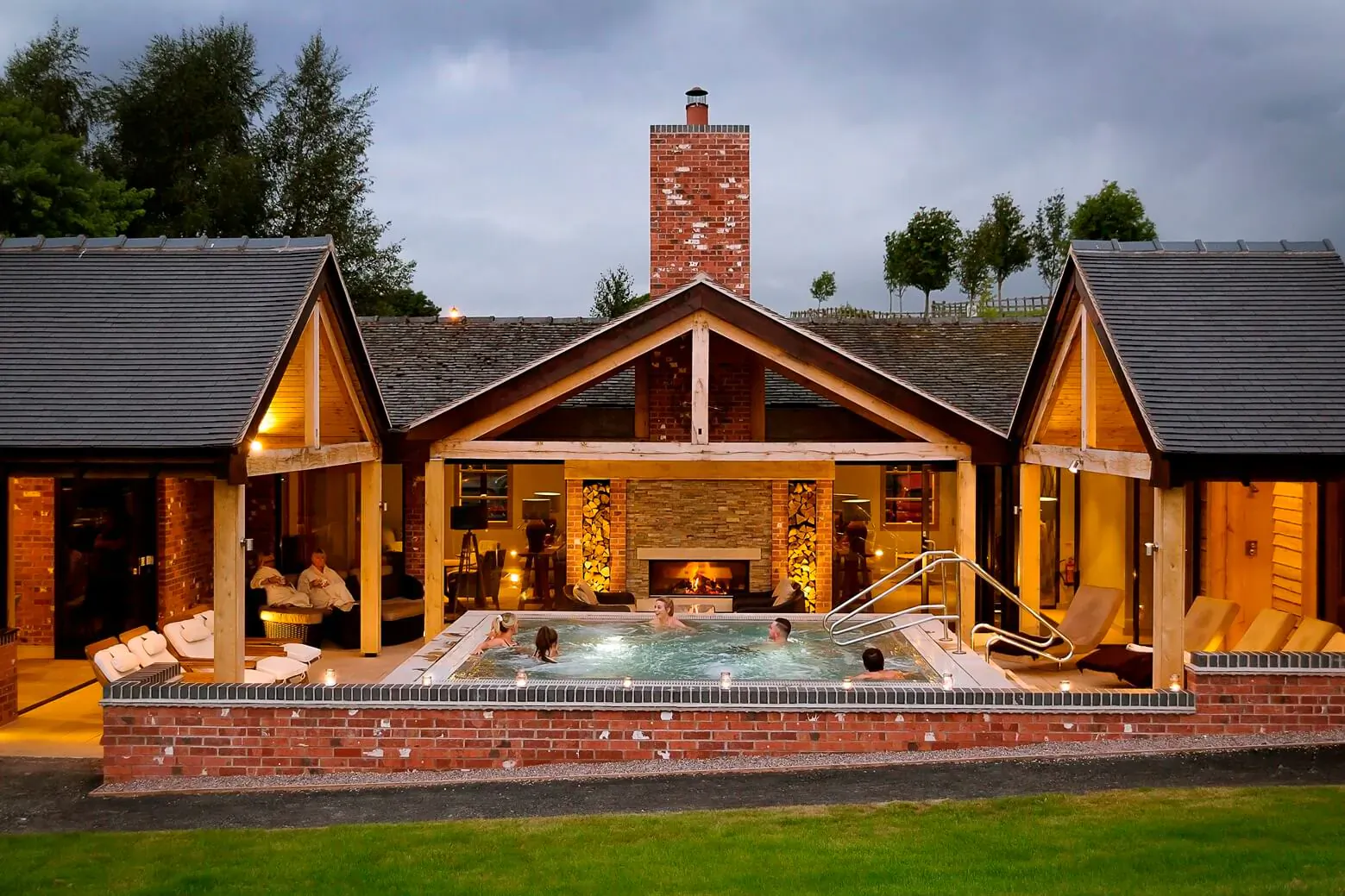 Twilight view of Moddershall Oaks spa: cosy wooden farmhouse with lit indoor pool, hot tub, loungers, fireplace, and people relaxing.