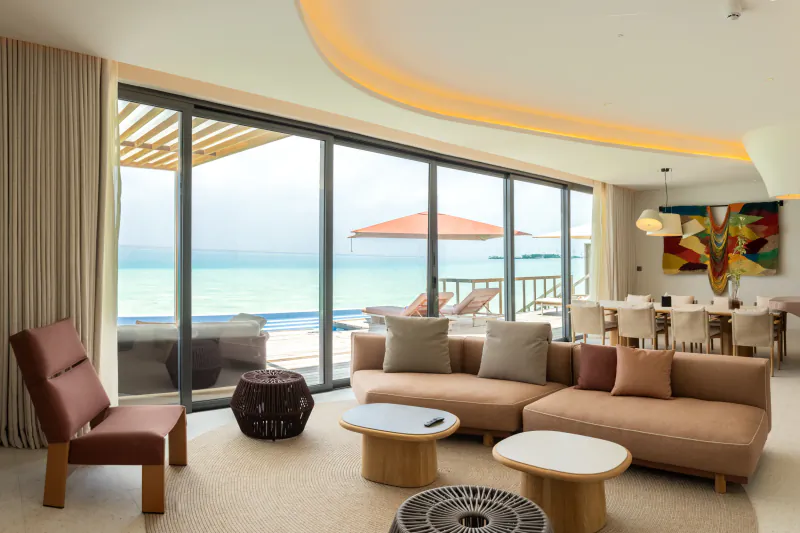 Luxurious modern living room in Crossroads Maldives villa with ocean views, beige sofas, dining table, and beach beyond glass doors.