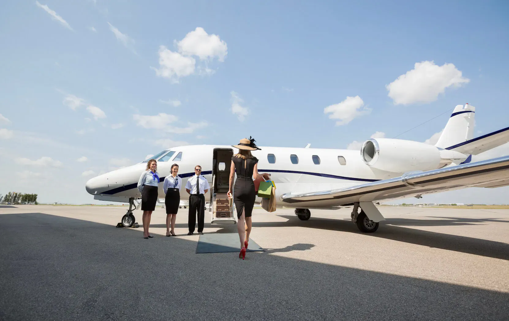 Elegant woman in black dress and hat with green bag walks toward white private jet, greeted by uniformed crew on tarmac under blue sky.