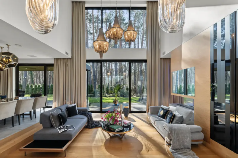 Spacious modern living room with gray L-shaped sofa, glass coffee table, flowers, dining area, and large windows overlooking forest.