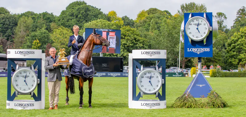 Two men in suits hold trophy atop chestnut horse on grass at Hickstead Derby, flanked by Longines clocks amid trees.