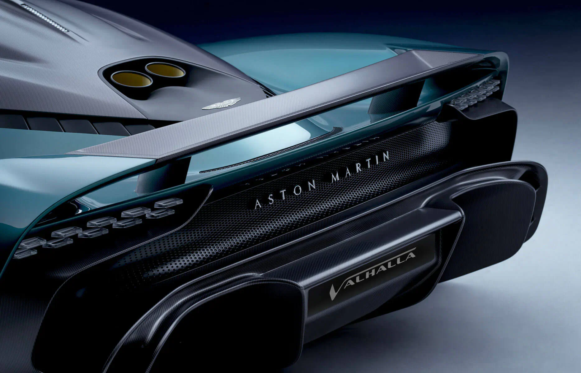 Rear view of teal Aston Martin Valhalla supercar with 'Aston Martin' and 'Valhalla' badges, yellow exhausts, and black diffuser.