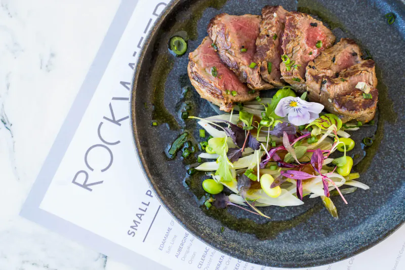 Sliced seared beef on dark plate with edamame, fennel, viola flowers, and green sauce at Rockwater restaurant.