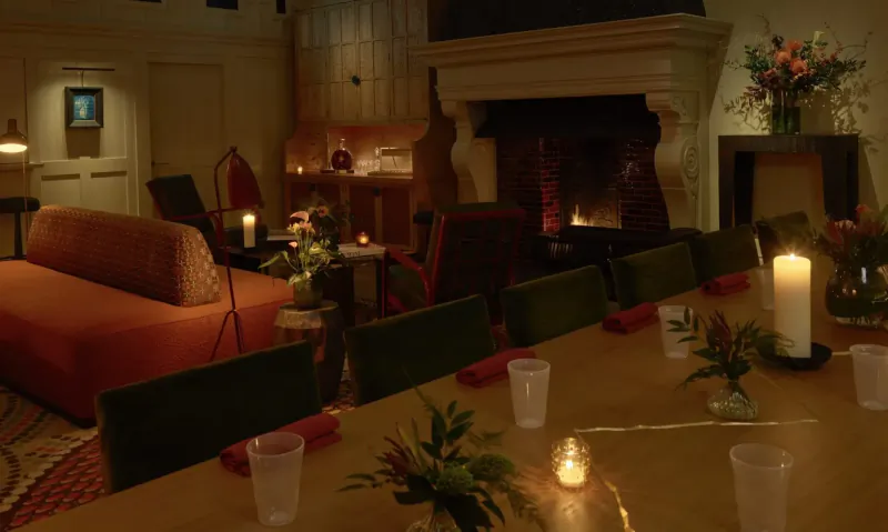 Cozy dining room with wooden table set for dinner, orange sofas, green chairs, flowers, candles, and warm fireplace glow.