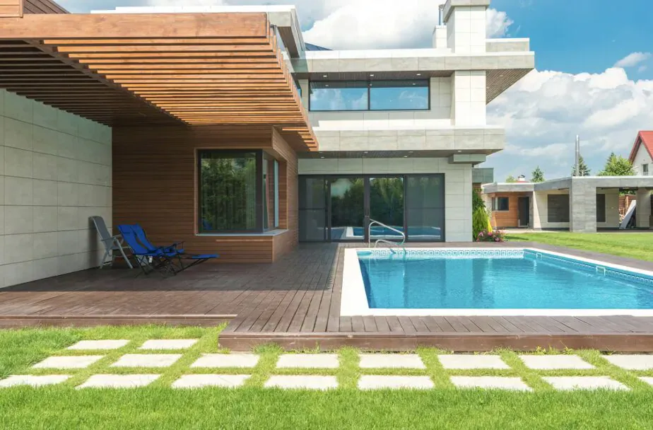 Modern luxury house with wooden deck, turquoise infinity pool, blue lounge chair, and lush green yard under blue sky.