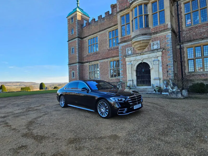 Black Mercedes S-Class sedan parked in front of a grand Tudor-style mansion with turrets and stone statues.