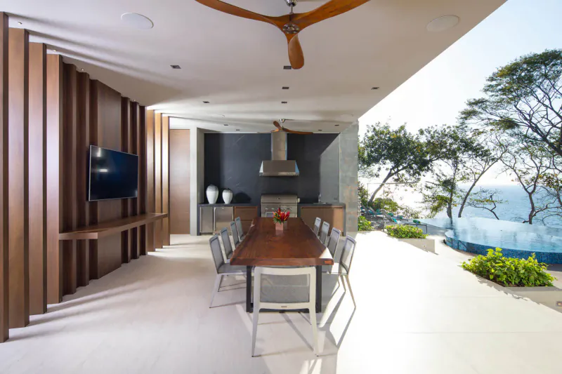 Modern outdoor kitchen and dining area with wooden table, chairs, TV, and ocean view from luxury home.