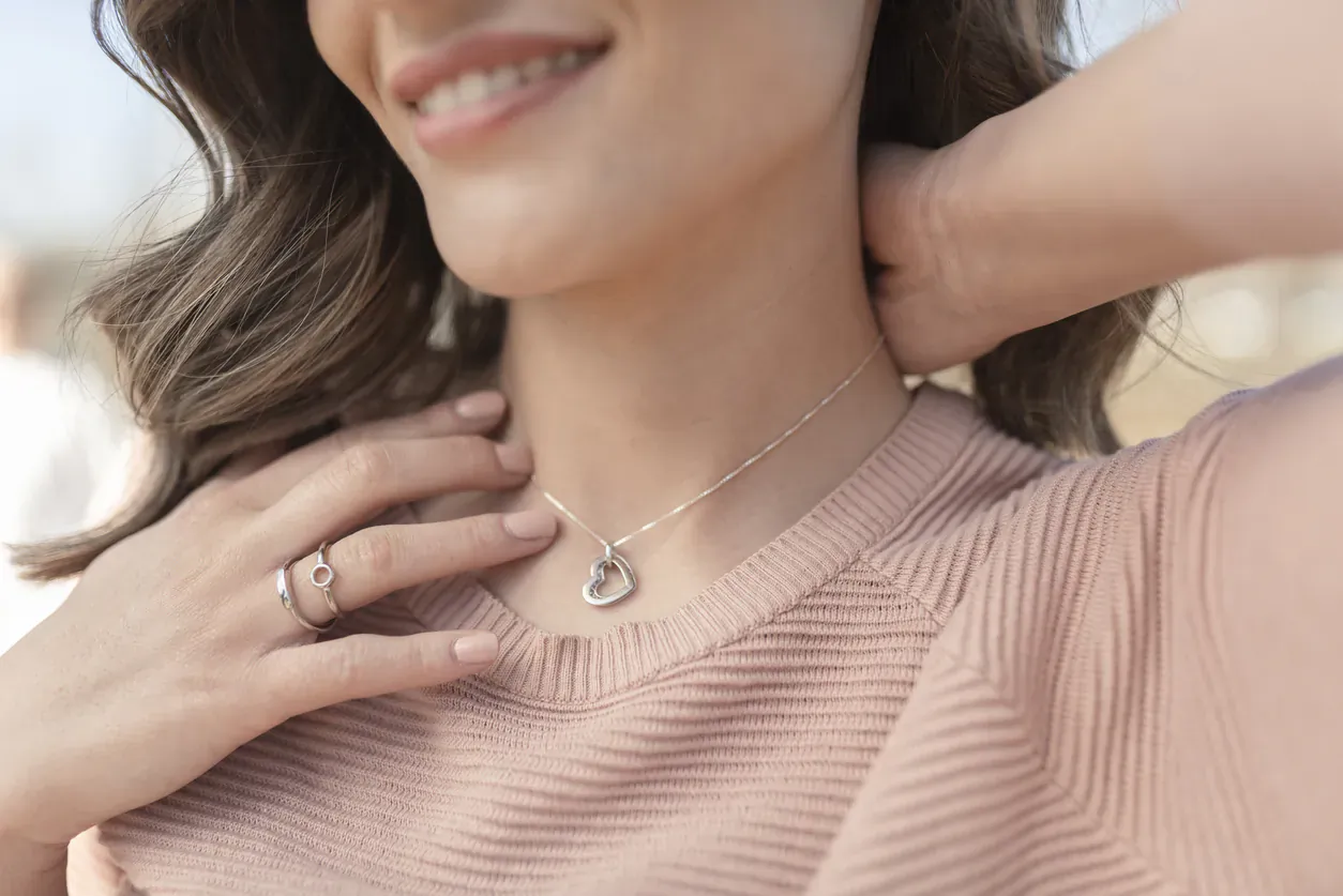 Smiling woman with wavy hair holding back her hair to showcase a heart pendant necklace on pink sweater
