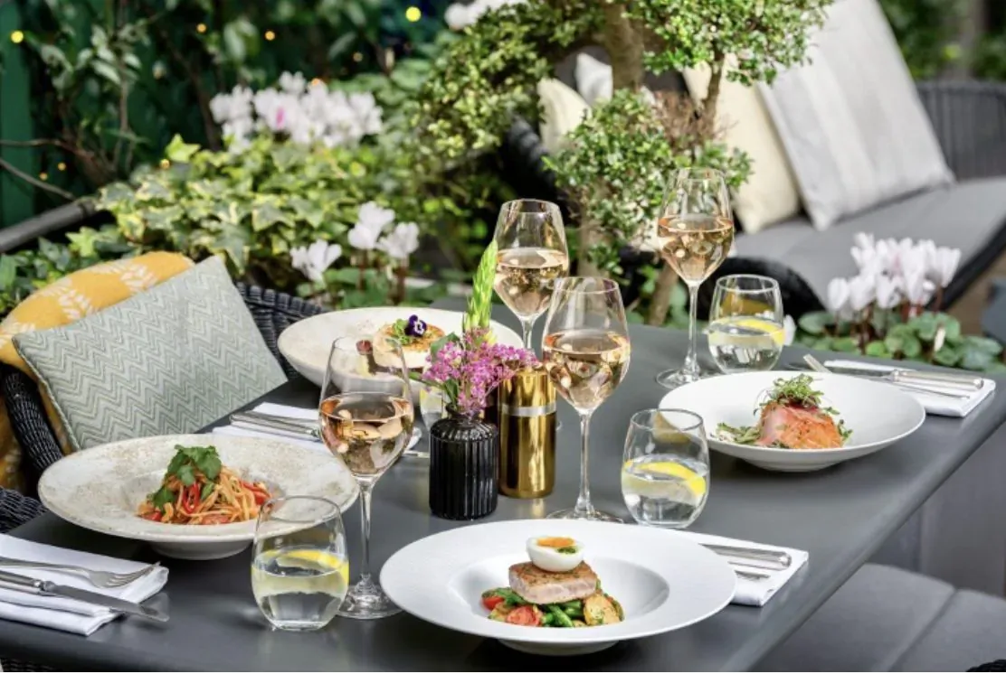 Elegant outdoor terrace table at The Sloane Club with rosé wine glasses, salmon, pasta, egg dish amid lush greenery and flowers.