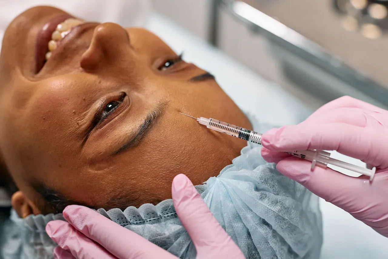 Close-up of Black woman smiling with eyes closed as gloved medical professional injects filler into her forehead in clinic.