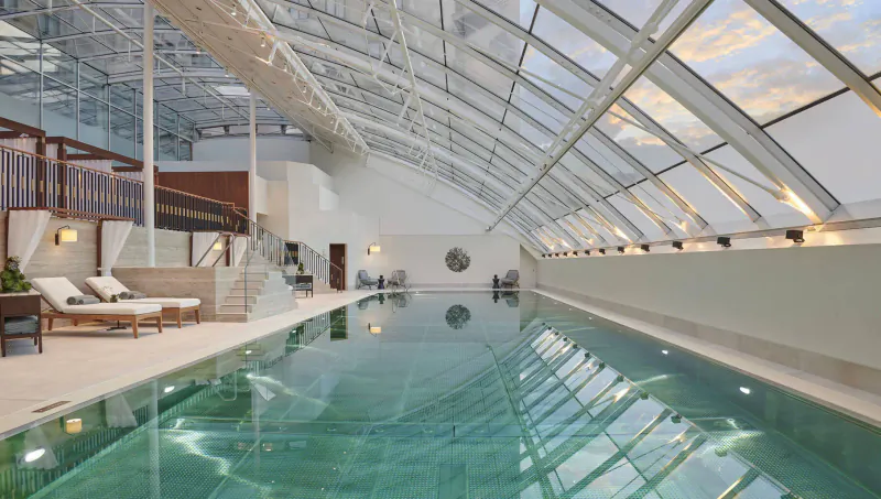Elegant indoor pool at Carlton Tower Jumeirah Hotel with white loungers, wooden accents, and expansive glass ceiling.